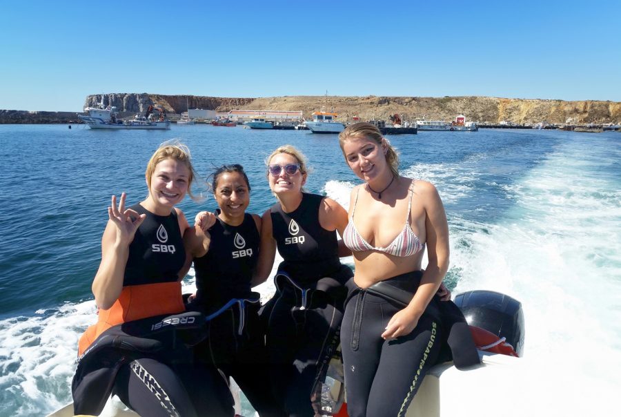 Four girls in wetsuits smile on a speedboat, with a rocky harbor and clear blue water behind them.