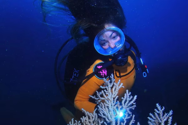 A scuba diver with a mask and orange suit floats beside white coral in deep blue water, with hair drifting upward.
